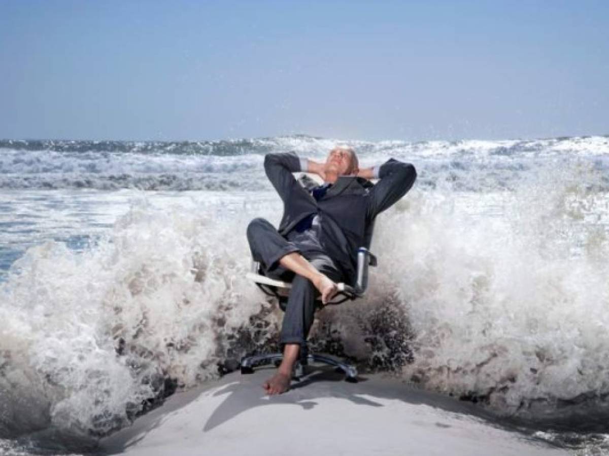 Senior business man sitting on office chair on beach, being splashed by waves