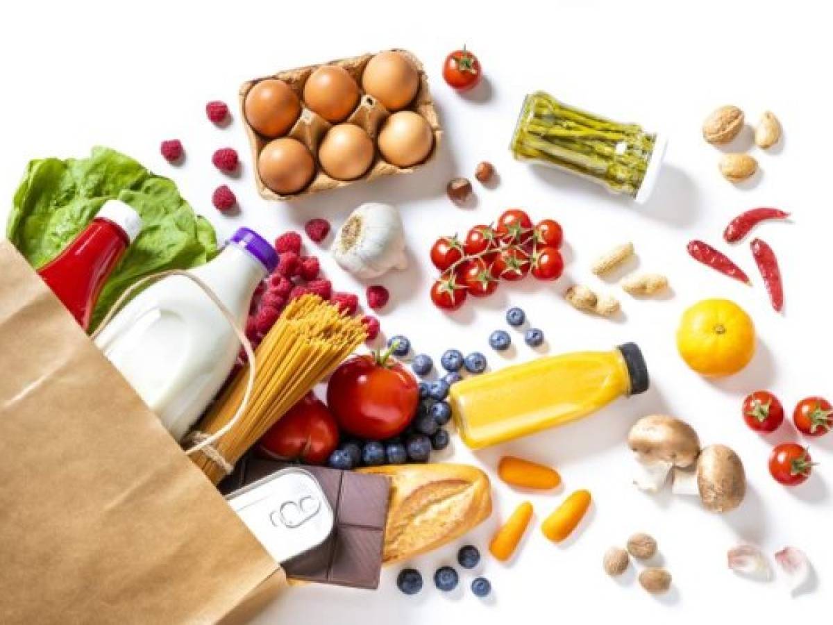 Top view of a paper bag full of canned food, fruits, vegetables, eggs, a milk bottle, berries, mushrooms, nuts, pasta, a chocolate bar and bread. The paper bag is laying on the white background and the food is coming out from it.