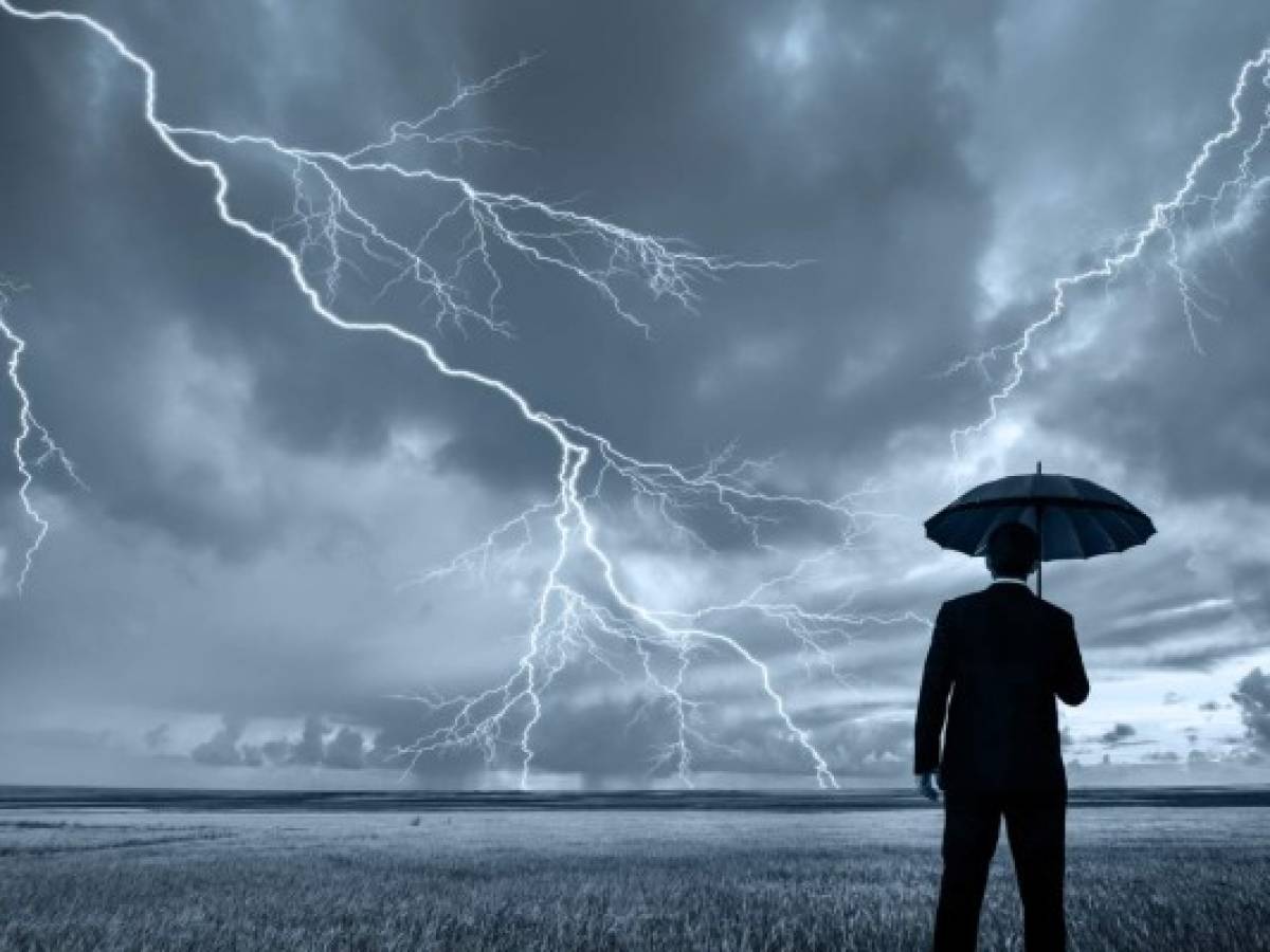 A rear view of a businessman in a suit, with an upheld umbrella, standing in a large field during a thunderstorm. Lightning is seen descending from a gray and cloudy sky.