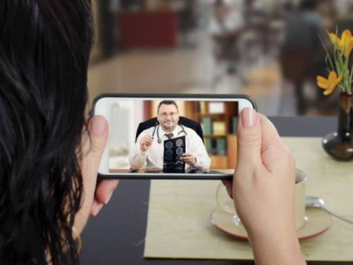Woman sits at a cafe table and communicates with telemedicine doctor by cellphone. In touchscreen, male physician reviewing brain x-ray image. Patient has ability to reach telemedicine specialist remotely by mobile application. Horizontal indoors back side shot on cafe blurred background Woman sits at a cafe table and communicates with telemedicine doctor by cellphone. In touchscreen, male physician reviewing brain x-ray image. Patient has ability to reach telemedicine specialist remotely by mobile application. Horizontal indoors back side shot on cafe blurred background