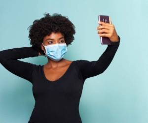 young woman with protection mask against corona virus using her mobile device to make a selfie wearing black shirt on blue background