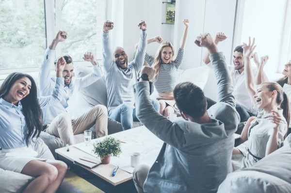 Group of young business people raising their arms and looking happy while sitting around the desk together