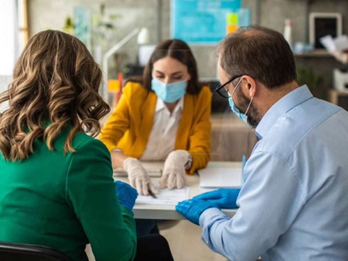 Mature adult couple sitting at desk with sneeze guard on it, talking with female bank manager and signing contract in bank, they are wearing protective face mask and surgical gloves for protection against virus