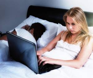 A handsome young couple lie in bed; she's intent on her laptop, either keeping up with social media, working late, or battling insomnia; he's fast asleep next to her.
