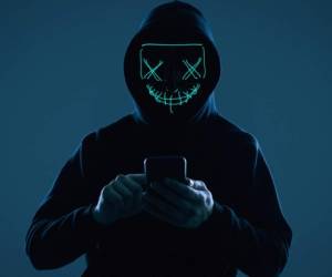 Portrait of an anonymous man in a black hoodie and neon mask hacking into a smartphone. Studio shot.