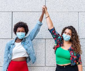 two young women friends, one white and one black, raise their fists together as a sign of sisterhood and protest against racism, new socialization with protective masks