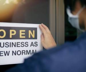 Reopening for business adapt to new normal in the novel Coronavirus COVID-19 pandemic. Rear view of business owner wearing medical mask placing open sign 'OPEN BUSINESS AS NEW NORMAL' on front door.