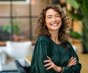 Portrait of young smiling woman looking at camera with crossed arms. Happy girl standing in creative office. Successful businesswoman standing in office with copy space.