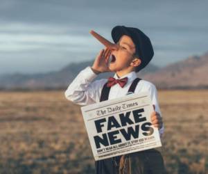 A news boy dressed in vintage knickers, newsboy hat and fake long Pinocchio nose stands with a fake newspaper in the middle of a field in Utah, USA. He is trying to sell you fake news.