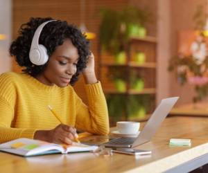Smiling black girl with wireless headset studying online, using laptop at cafe, taking notes, copy space