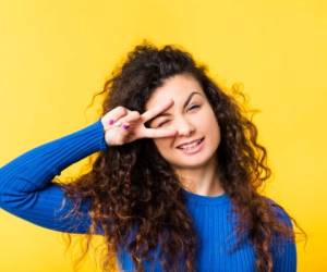 Portrait of cheerful brunette girl with curly hair. Funny emotional lady winking eye through fingers in victory gesture, smiling.