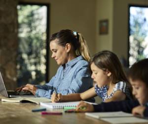 Confident businesswoman using laptop at table. Mother is sitting by children doing homework. They are at home.