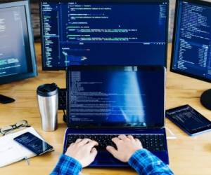 Multiple computer screens with program code on office table. Programmer, developer hands are typing on keyboard. Devices for working, tablet, smartphone, virtual glasses, notebook on geek workplace.