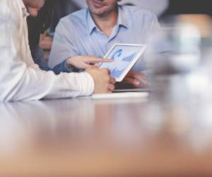 Business people working on financial data on a digital tablet. One looks like the manager working with employees, or a financial advisor. It may also be a training session. All are dressed in business casual clothing.