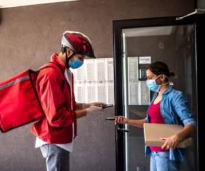 Pizza delivery person wearing red jacket, cycling helmet, protective face mask and surgical gloves, scanning bar code on customers smart phone and making a quick and easy contactless payment