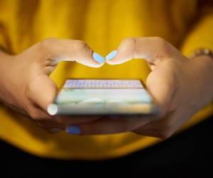 Young woman using cell phone to send text message on social network at night. Closeup of hands with computer laptop in background