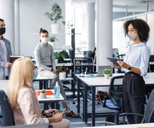 Corporate meeting and group work in modern company in office interior. African american woman manager in protective mask holding tablet, talking to workers keeping social distance during epidemic