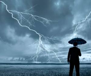 A rear view of a businessman in a suit, with an upheld umbrella, standing in a large field during a thunderstorm. Lightning is seen descending from a gray and cloudy sky.