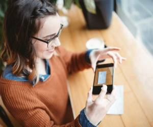 A smiling young woman takes a picture with her smart phone of a check or paycheck for digital electronic depositing, also known as 'Remote Deposit Capture'. She sits in a coffee shop, enjoying an espresso latte. Bright sunlight shines in the window. Horizontal image.