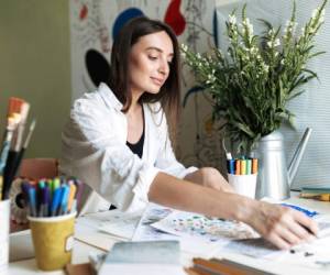 Young beautiful painter sitting at the desk with pictures dreamily working at cozy home