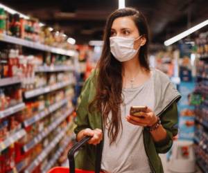 Image of a young woman in the supermarket during the coronavirus pandemic. She is checking the list of things to buy on her smartphone app.