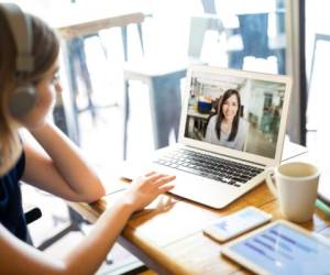 Woman wearing headphones and participating in a video conference call on a laptop while telecommuting from a cafe