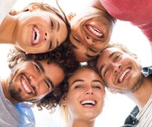 Directly below shot of young friends forming huddle. Low angle view of girls and guys with their head forming a circle. Portrait of young people looking at camera. Friendship and unity concept.