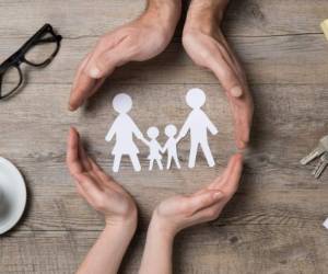 Close up of female and male hands protecting a paper chain family. Top view of two hands form a circle around white paper chain family on wooden table. Family care, insurance and helping hand concept.