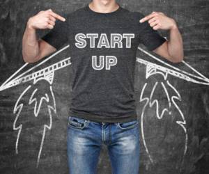 A businessman is pointing out in the chest where is a word collocation ' start up '. A concept of the starting up new business. Drawn wings. Black chalk board on the background.