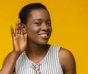 Curious interested african american woman holding hand near ear and trying to hear information, yellow studio background