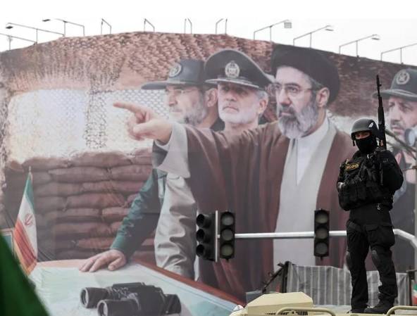 Fotografía de archivo de un miembro de las fuerzas de seguridad iraníes frente a una valla en la que aparecen el líder supremo iraní, Mojtaba Jamenei, y varios mandos militares iraníes. EFE/EPA/ABEDIN TAHERKENAREH