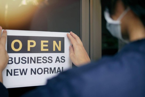 Reopening for business adapt to new normal in the novel Coronavirus COVID-19 pandemic. Rear view of business owner wearing medical mask placing open sign 'OPEN BUSINESS AS NEW NORMAL' on front door.