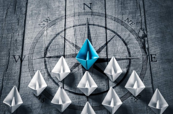 Blue Paper Boat Leading A Fleet Of Small White Boats With Compass Icon On Wooden Table - Leadership Concept