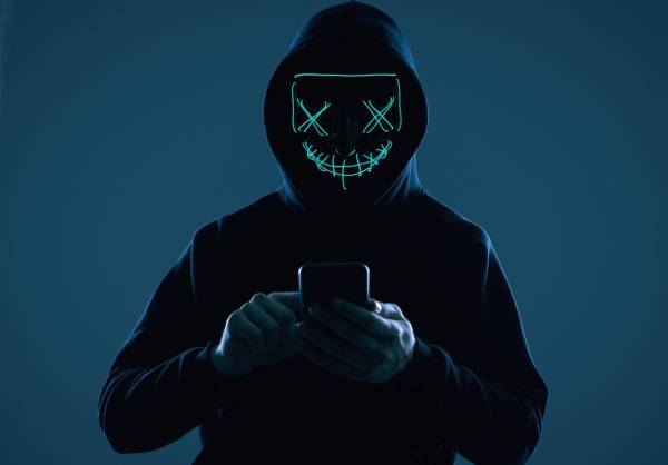 Portrait of an anonymous man in a black hoodie and neon mask hacking into a smartphone. Studio shot.