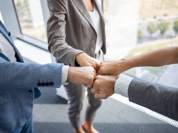 Hands together in fist bump for agreement, four business people standing by the office window on high floor