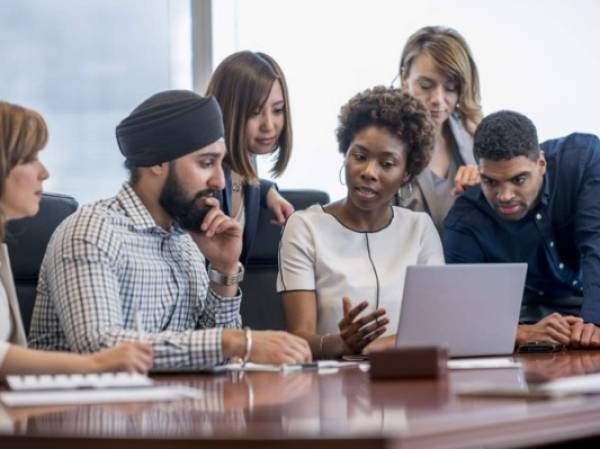 A diverse group of business people gather around a laptop in a modern office and discuss what they see.