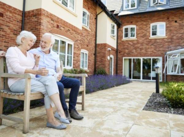 Retired Couple Sitting On Bench With Hot Drink In Assisted Living Facility