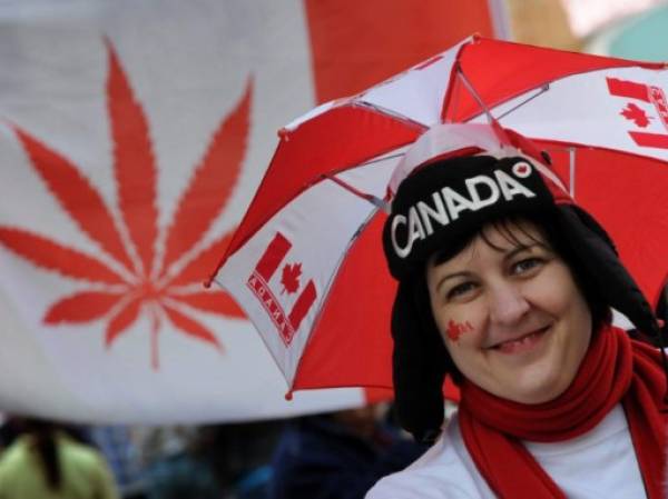 A Canadian fan wears an umbrella hat in front of a Cannabis flag in Vancouver during the Winter Olympics on February 25, 2010. AFP PHOTO / Mark RALSTON (Photo by MARK RALSTON / AFP)